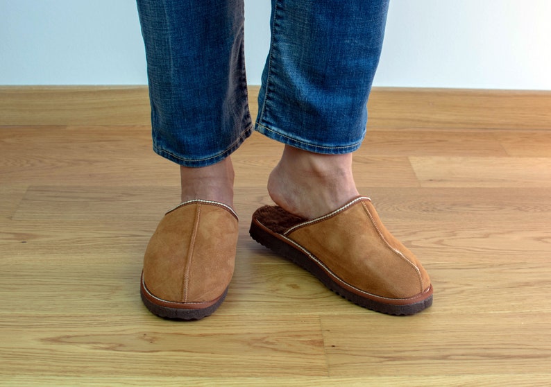 May include: A pair of brown suede slippers with a white trim. The slippers are worn on a wooden floor.