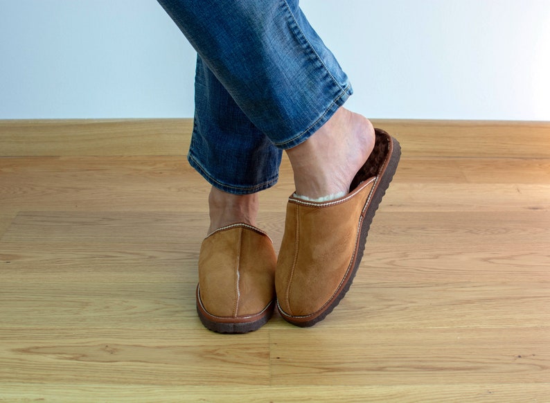 May include: A pair of brown suede slippers with a white sheepskin lining. The slippers are worn by a person wearing blue jeans.