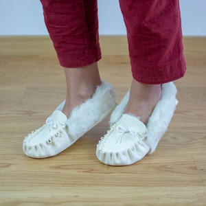 本物のムートンレザーで作られた女性用モカシン。内側は柔らかい毛皮、上部にはかわいいリボンが付いています。手作りの白いホームシューズ。彼女へのプレゼントに最適です。