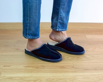 May include: A pair of dark blue suede slippers with white stitching. The slippers are being worn by someone wearing blue jeans.