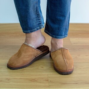 May include: A pair of brown suede slippers with a white trim. The slippers are worn by a person wearing blue jeans.