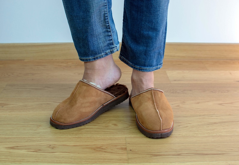 May include: A pair of brown suede slippers with a white trim. The slippers are worn by a person wearing blue jeans.