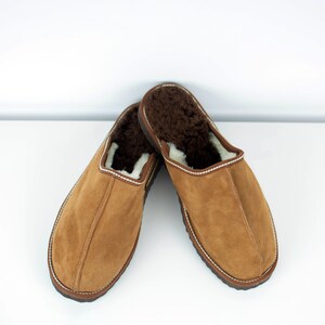 May include: A pair of brown suede slippers with a white sheepskin lining. The slippers have a closed toe and a rounded shape.