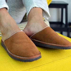 May include: A pair of brown suede slippers with white stitching and a dark brown sole. The slippers are being worn by someone sitting on a yellow sofa.