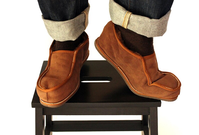 Pode incluir: Chinelos de camur&ccedil;a castanhos com design de mocassim, com costuras brancas e acabamento contrastante. Usados com jeans azul escuro com bainhas dobradas. Os chinelos est&atilde;o sobre um pequeno banco de madeira preto.