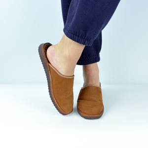 May include: A pair of brown leather slippers with a suede upper and a stitched seam around the edge. The slippers are being worn by someone wearing blue trousers.
