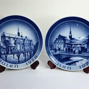 May include: Two blue and white decorative plates with church scenes. The plates, labeled "Julen" and dated 1978 and 1979, feature detailed illustrations of buildings and trees. The plates are displayed on small wooden stands.
