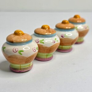 May include: Four small ceramic jars with lids, arranged in a diagonal line. Each jar features a rounded body with a tan upper section, a white middle band decorated with small pink swirls and green leaves, and a light green base with pink trim.