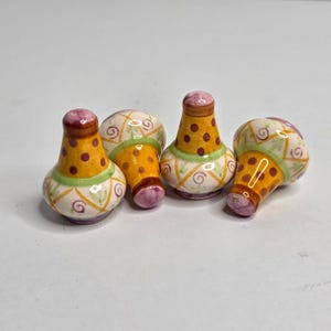 May include: Four decorative ceramic salt and pepper shakers. Each shaker has a bulbous base, a cylindrical body with a yellow polka dot pattern, and a purple top. The shakers are decorated with green and purple accents.