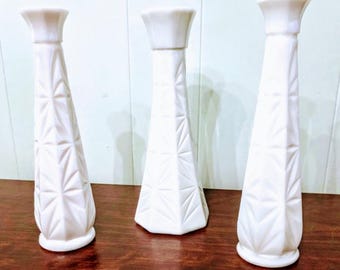 May include: Three white, vintage-style vases with geometric patterns. The vases have a flared top and a faceted, tapered body. They are displayed on a dark wooden surface.