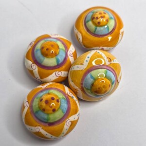 May include: Four round ceramic knobs featuring a glossy finish with a vibrant yellow base color. Each knob displays a central raised dome with brown polka dots, surrounded by concentric rings of light blue, green, and purple, with white swirls.