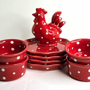 May include: A red ceramic rooster figurine with white polka dots sits atop a stack of matching plates. Two stacks of red bowls with white polka dots are on either side. This set is perfect for kitchen decor.