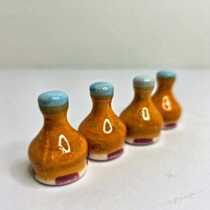 May include: Four small, bell-shaped ceramic objects with a glossy finish. Each piece features a rounded orange body, a light blue top, and a small purple rectangular accent at the base. The surface is decorated with subtle, scattered green and brown markings.