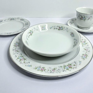 May include: A white porcelain dinnerware set with a floral pattern in blue, pink, and green. The set includes a dinner plate, a salad plate, a bowl, a saucer, and a teacup. The edges of the plates and cup are trimmed with a silver band.