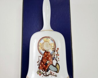 May include: White ceramic bell with a long handle and a detailed illustration of a child with a halo, wearing a red coat and blue pants. The bell has a gold trim and is set against a dark blue background.