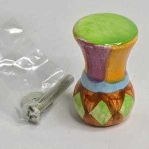 May include: A ceramic cabinet knob with a glossy finish, featuring a bright green top and a body painted with vertical stripes of purple, orange, and light blue, ending in a base with a brown and green diamond pattern, accompanied by a metal bolt.