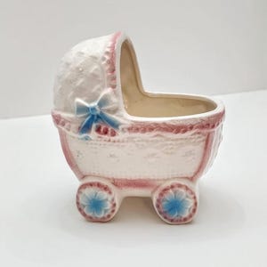 May include: A ceramic baby carriage planter in white, pink, and blue. The carriage features a decorative bow, floral accents on the wheels, and a lace-like pattern. The interior is beige.