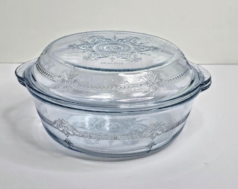 May include: A round, light blue glass casserole dish with a matching lid. The exterior features an embossed, intricate scrollwork pattern along the rim and a central starburst design on the lid, complete with two integrated side handles.