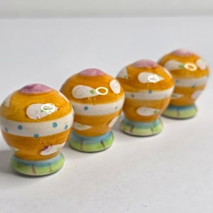 May include: Four rounded ceramic knobs featuring a glossy finish with horizontal yellow stripes, small blue dots, and white cloud-like shapes. Each knob has a pink top and a light green base, providing a colorful and smooth surface for furniture.