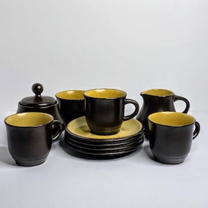 May include: A ceramic tea set featuring dark brown exteriors and speckled yellow interiors. The set includes four cups, four saucers, a lidded sugar bowl, and a creamer pitcher. Each piece has a smooth, rounded shape with a consistent color scheme.