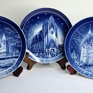May include: Three blue and white decorative plates with church scenes. Each plate features a different church design, with the year and the word "Julen" printed on the bottom. The plates are displayed on wooden stands.