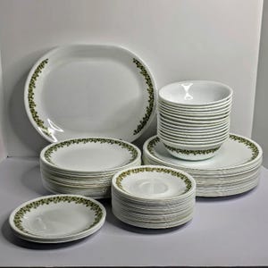 May include: A white dinnerware set featuring a green floral pattern along the edges. The collection includes a large oval platter, stacks of round dinner plates, smaller side plates, and a stack of deep bowls, all with a consistent green leaf design.