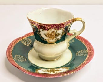 May include: A vintage teacup and saucer set. The cup and saucer feature a teal and cream color scheme with gold and maroon accents. The cup has a curved handle and a decorative floral design.