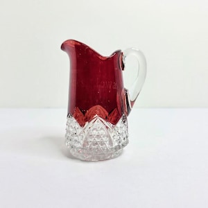 May include: A small, ruby red glass pitcher with a clear handle. The top portion is a deep red, while the bottom has a clear, cut-glass design. The word "Iowa" is faintly visible on the red portion. A decorative piece for serving.