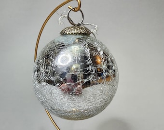 May include: A silver crackle glass Christmas ornament with a gold-colored metal cap and a silver ribbon. The ornament is suspended from a gold-colored metal stand with a wooden base. The ornament is round and reflective.