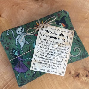 May include: A green pouch with a whimsical witch and cat design, tied with twine and a tag that reads "little bundle of everyday magic." The pouch is decorated with illustrations of witches, cats, and floral elements.