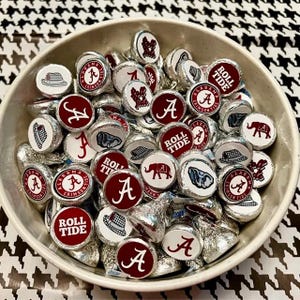 May include: A bowl filled with silver and red round sweets with various designs, including the words "Roll Tide", "Alabama Crimson Tide", and the letter "A".