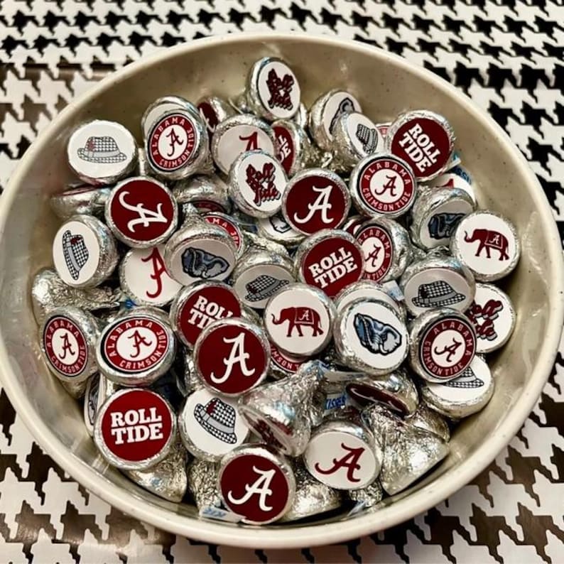 May include: A bowl filled with silver and red round sweets with various designs, including the words "Roll Tide", "Alabama Crimson Tide", and the letter "A".