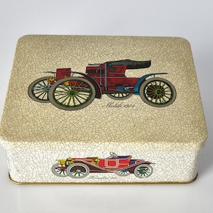 May include: A vintage tin box with a cream background and a crackled finish. The box features two illustrations of antique cars, one red and one gold, with the text "Model 1901" and "Manufactured 1903".