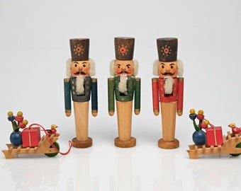 May include: Three wooden nutcracker figurines with painted faces and hats, standing in a row. Each nutcracker is wearing a different colored uniform: blue, green, and red. Two small wooden toy carts with red boxes and yellow flowers are on either side of the nutcrackers.