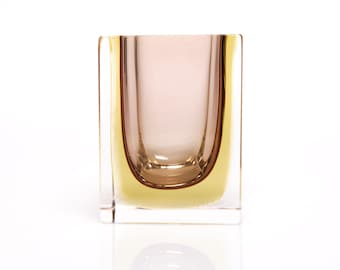 Puede incluir: Un jarrón de vidrio transparente con forma cuadrada y un diseño único en capas. El jarrón tiene un interior marrón claro y un exterior amarillo.