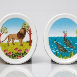 May include: Two white ceramic plates with a decorative border. The plate on the left features a brown lion with a red mane standing in a green field with pink flowers. The plate on the right features two blue fish with pink flowers on top of their heads swimming in a blue ocean with green seaweed.