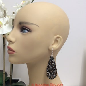 Peut inclure: Une paire de longues boucles d'oreilles en forme de goutte avec un motif de perles noires, blanches et dor&eacute;es. Les boucles d'oreilles sont suspendues &agrave; l'oreille d'un mannequin.