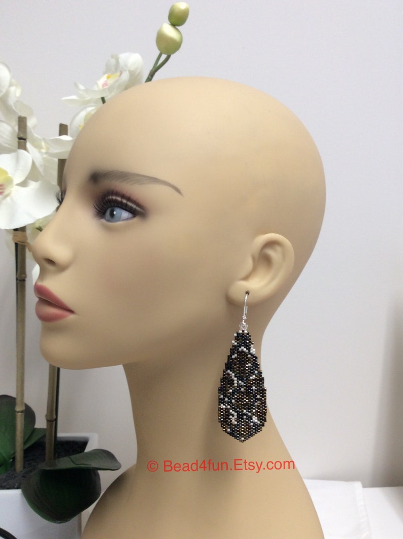 Peut inclure: Une paire de longues boucles d'oreilles en forme de goutte avec un motif de perles noires, blanches et dor&eacute;es. Les boucles d'oreilles sont suspendues &agrave; l'oreille d'un mannequin.