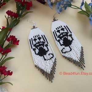 May include: A pair of long, white beaded earrings with a black and white geometric design. The earrings have a silver-colored fringe at the bottom.