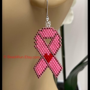 May include: Pink beaded ribbon earrings with a red heart-shaped accent. The earrings are made with small beads and have silver-colored ear wires. The earrings are a symbol of breast cancer awareness.