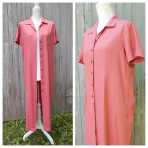 May include: A long, coral-pink button-down dress with short sleeves and a classic collar. The garment features a full-length front button placket with matching buttons, a straight hemline, and a simple, unadorned design displayed on a mannequin.