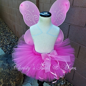 May include: Pink tulle tutu skirt with a pink ribbon bow and matching pink fairy wings with silver glitter accents.  "Christy's Tutus & More" is written on the skirt.