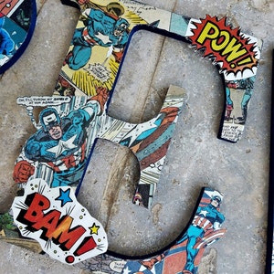May include: A wooden letter E covered in comic book pages featuring Captain America. The letter is painted blue and has a red, yellow, and white POW! comic book speech bubble.