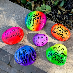 Può includere: Una collezione di sei pietre lisce dipinte a mano con vivaci sfumature di colore e scritte nere. Le pietre riportano le frasi "YOU GOT THIS", "love yourself", "Smile and SHINE", "BE KIND", "You Matter" e una faccina sorridente di colore viola.