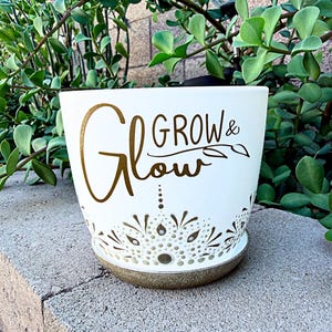 Puede incluir: Una maceta de cerámica blanca con las palabras "Grow & Glow" en escritura dorada. La maceta tiene un diseño decorativo perforado alrededor de la base y se asienta sobre un platillo a juego. El follaje verde es visible en el fondo.