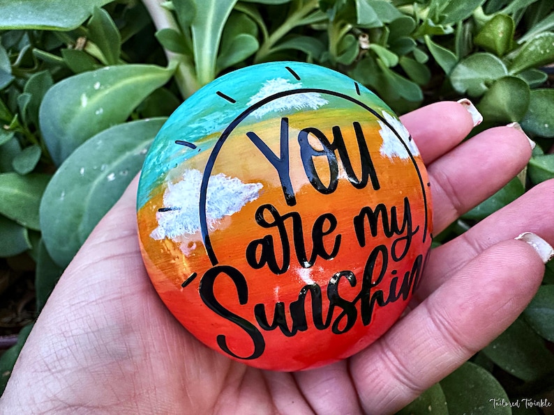 Op de afbeelding: Een beschilderde steen met een regenboog-zonsondergangontwerp en de woorden "You are my sunshine" in zwarte letters.