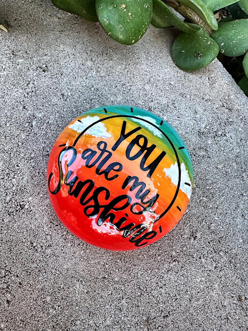 Op de afbeelding: Een beschilderde steen met een zonsondergangsc&egrave;ne en de woorden "You are my sunshine" in zwarte letters.