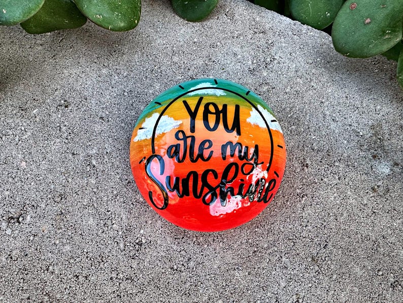 Op de afbeelding: Een beschilderde steen met een regenboogkleurige achtergrond en een zon met wolken. De tekst "You are my Sunshine" staat in het zwart op de steen.