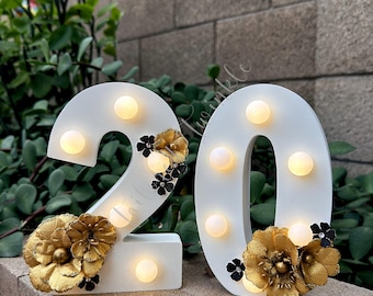 May include: Two white number signs, 2 and 0, featuring integrated warm white LED bulbs. Each number is decorated with metallic gold fabric flowers and small black floral accents. The numbers are freestanding and display a smooth matte finish.
