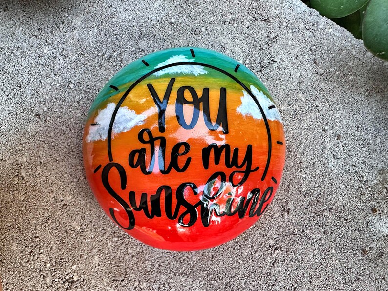 Op de afbeelding: Een beschilderde steen met een regenboog-zonsondergangontwerp en de woorden "You are my Sunshine" in zwarte letters.